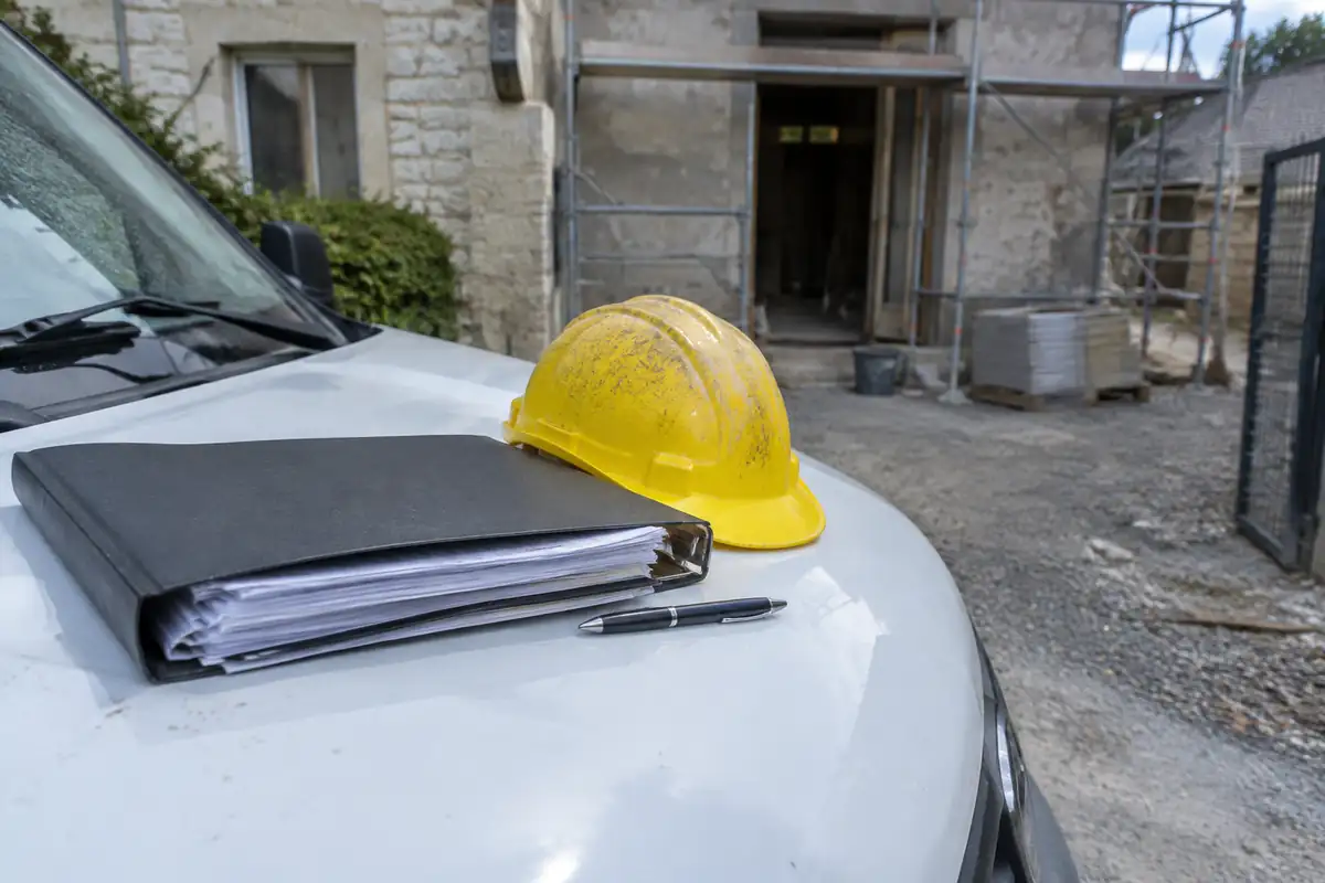 Dossier de rapports de vérification et casque jaune posés sur le capot d'une camionnette devant un chantier