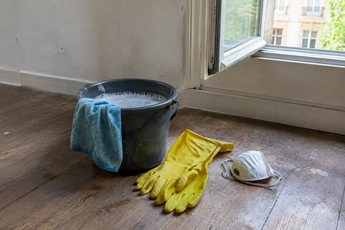 Seau d'eau savonneuse avec une microfibre, gants jaunes et masque posés sur un parquet devant une fenêtre ouverte